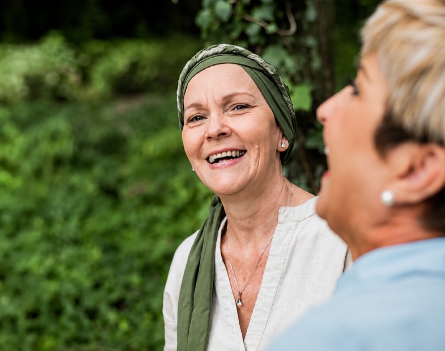 Duas mulheres conversando e rindo ao ar livre; uma delas usa lenço na cabeça